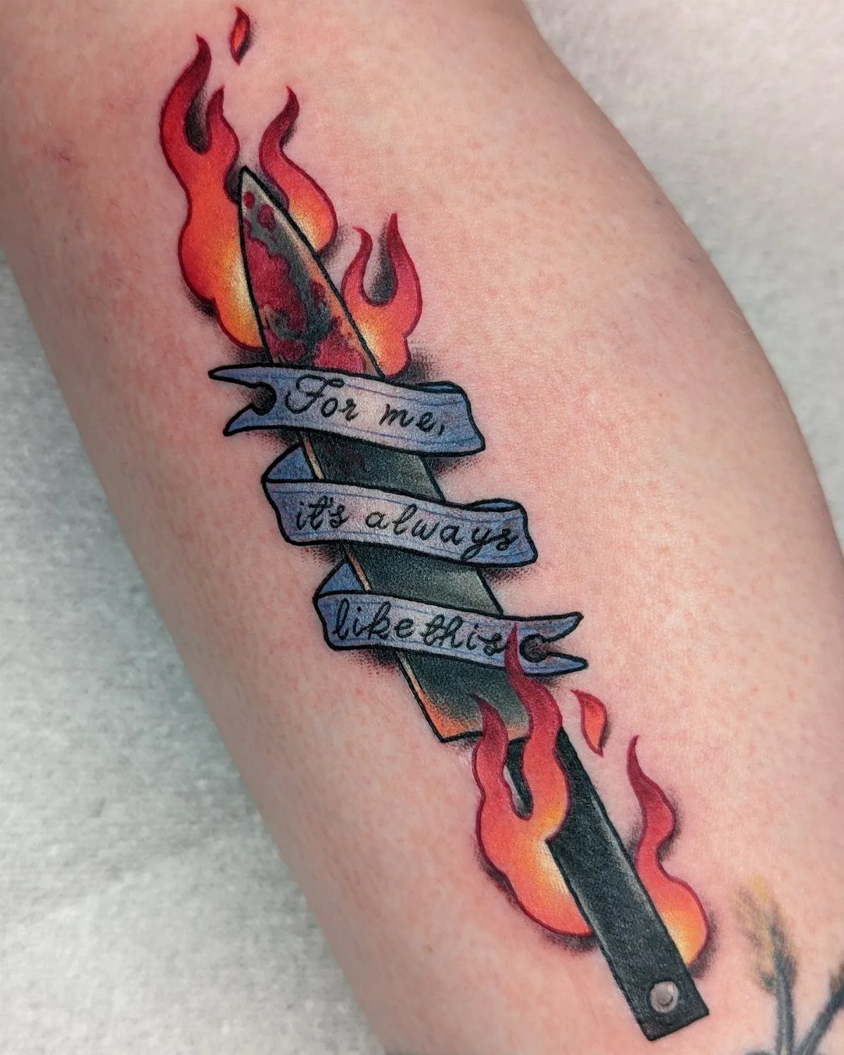 Knife tattoo with red flames and a blue ribbon with text on skin