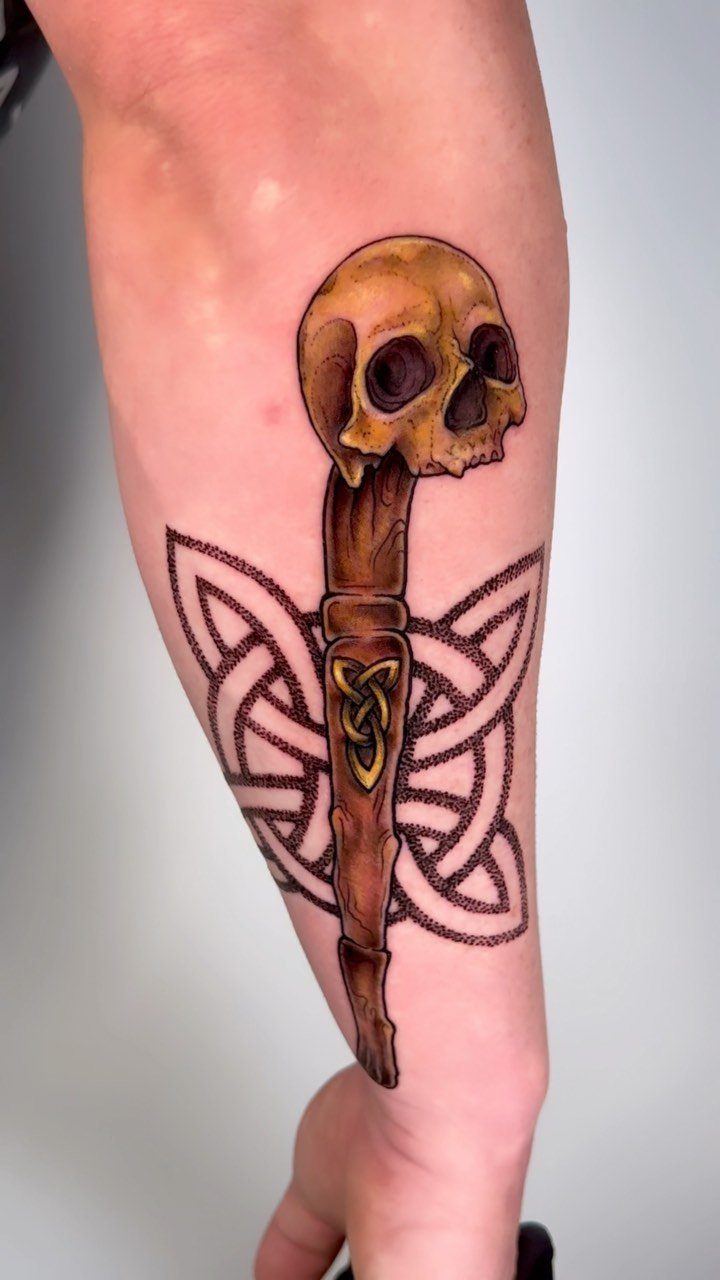 Skull on a wooden staff with Celtic knot patterns tattooed on a forearm.