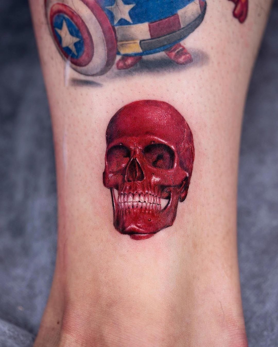Red skull tattoo on an ankle with a Captain America shield tattoo above