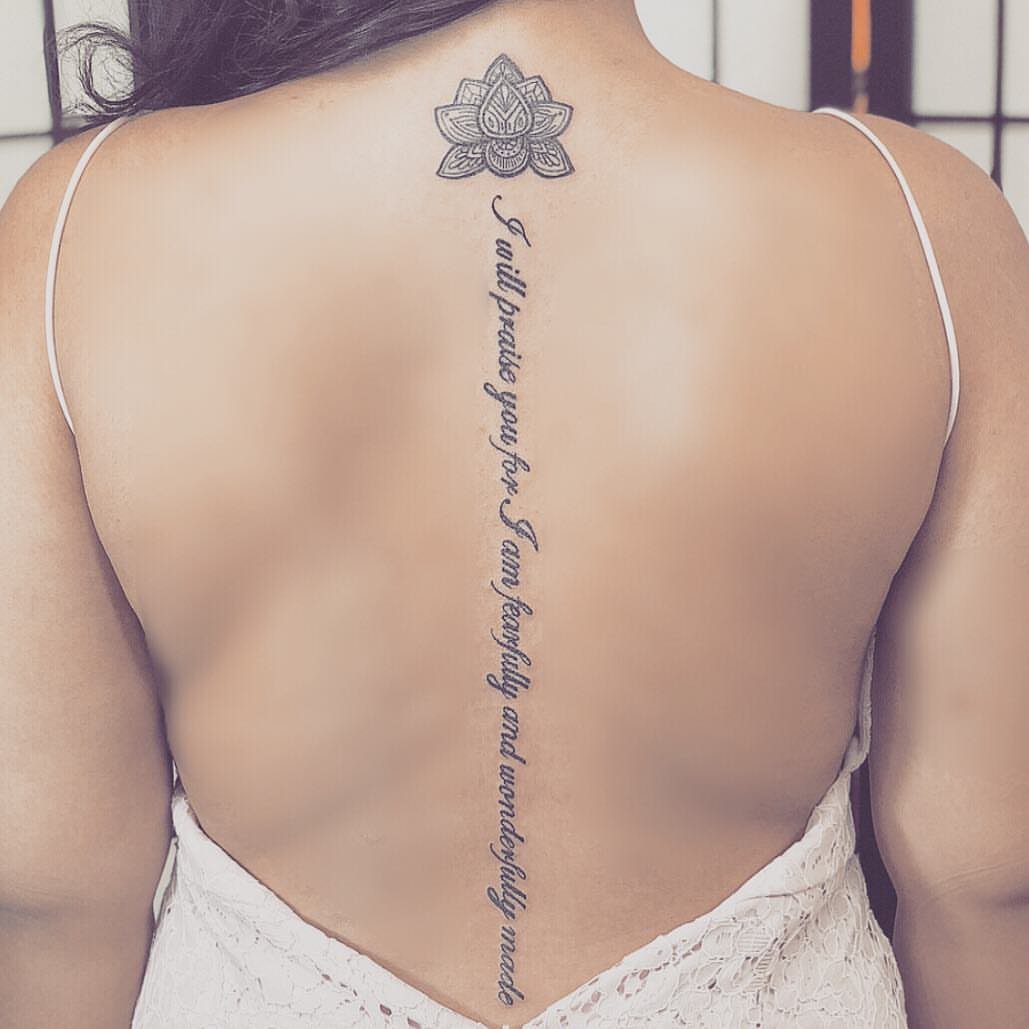 Lotus flower and script sentence tattoo running vertically down a woman's spine.