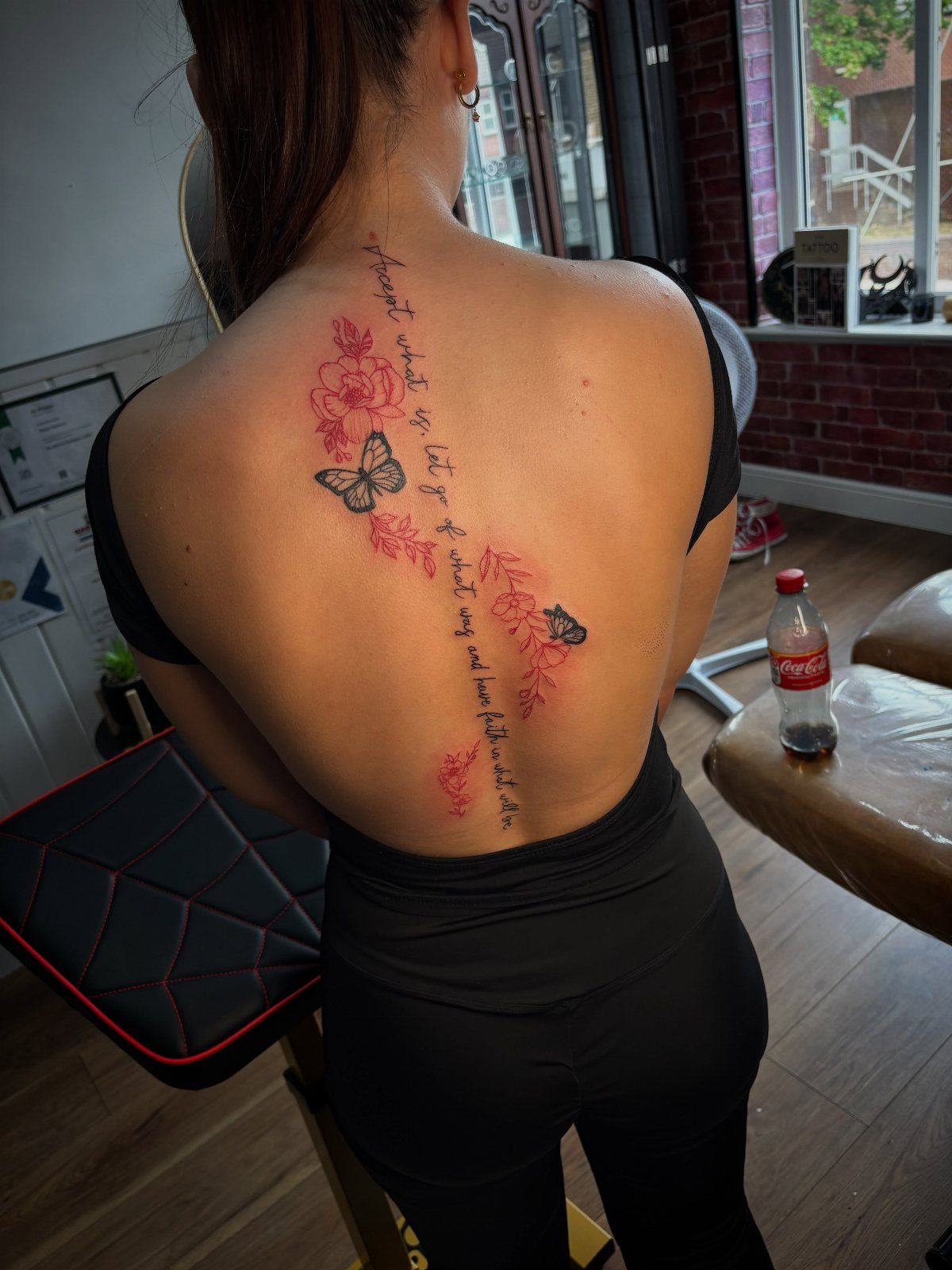 Woman's back with a red spinal tattoo featuring butterflies, flowers, and cursive text.