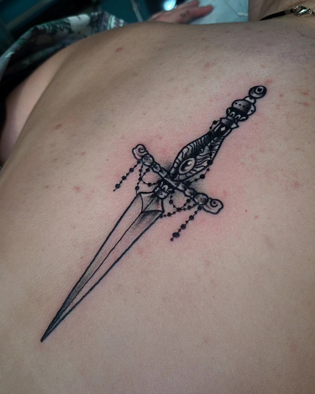 Ornate black ink dagger tattoo on a person's back with detailed hilt.