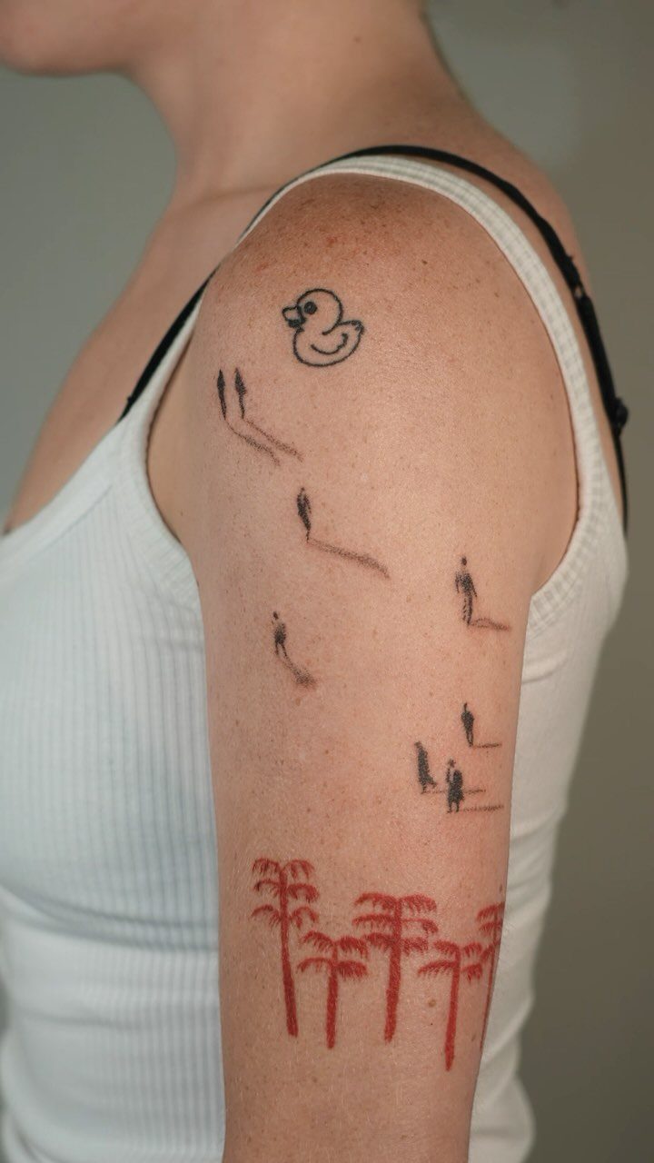 Hand-poked tattoos of a duck, tiny figures, and red palm trees on an arm.