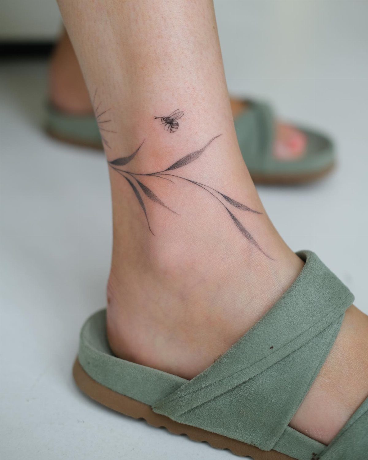 Minimalist black ink tattoo of a leafy branch and bee on an ankle.