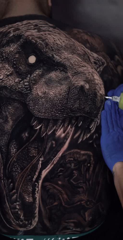 Realistic T-Rex tattoo on a person's back being worked on by an artist.