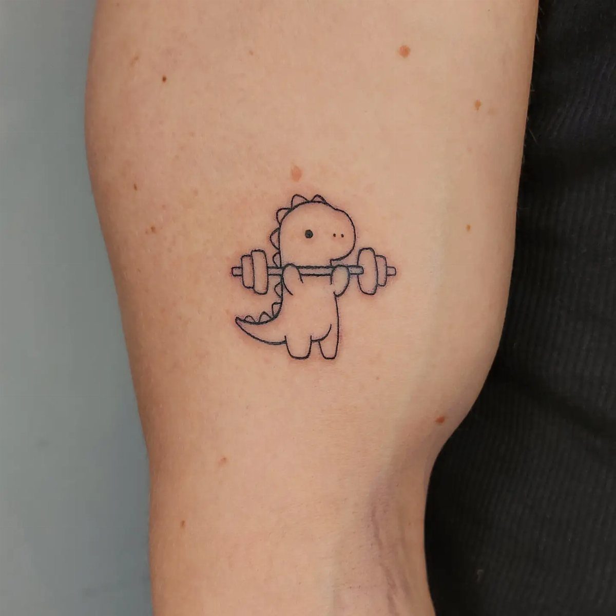 Small minimalist T-Rex tattoo lifting a barbell on a person's upper arm.