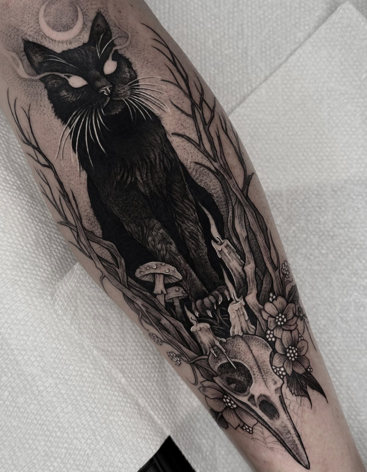 Black cat tattoo with a crescent moon, bird skull, and spooky forest elements.