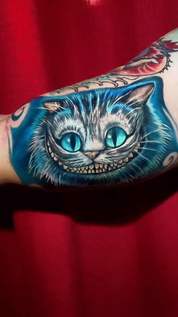 Cheshire Cat tattoo with a wide grin and glowing blue eyes on an arm.