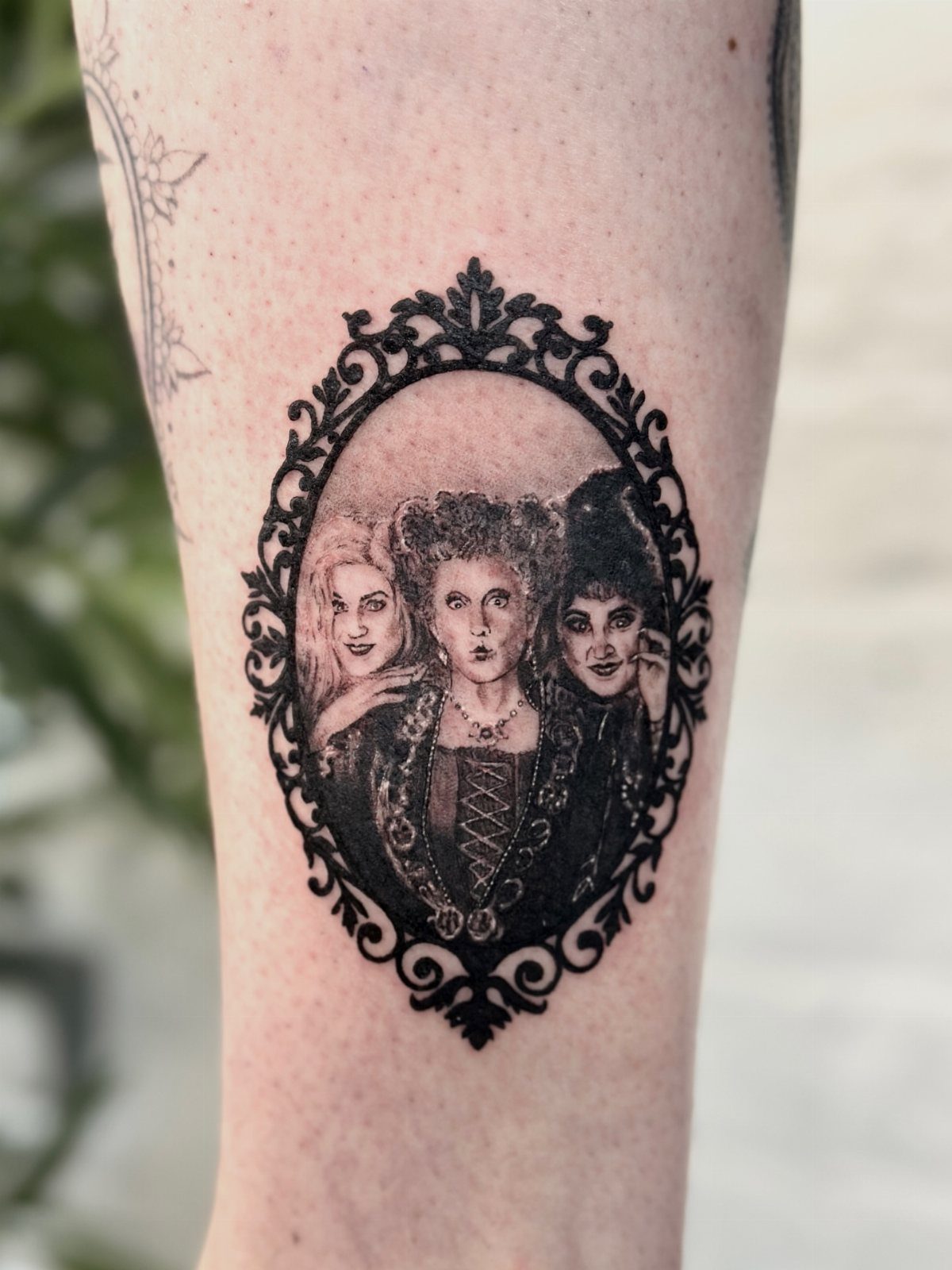 Tattoo of the three Sanderson sisters inside an ornate oval frame on skin.