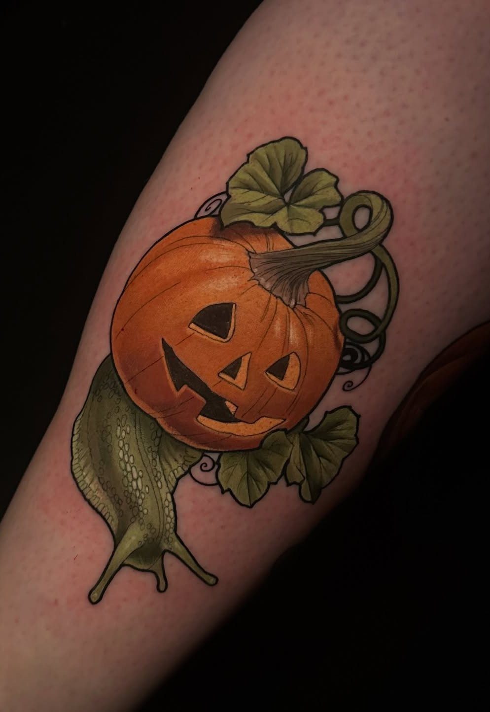Tattoo of a green snail with a carved pumpkin jack-o'-lantern shell and leaves.