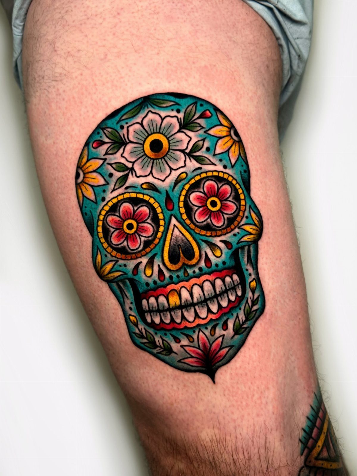 Sugar skull tattoo with colorful flowers and teal skin on a person's leg.