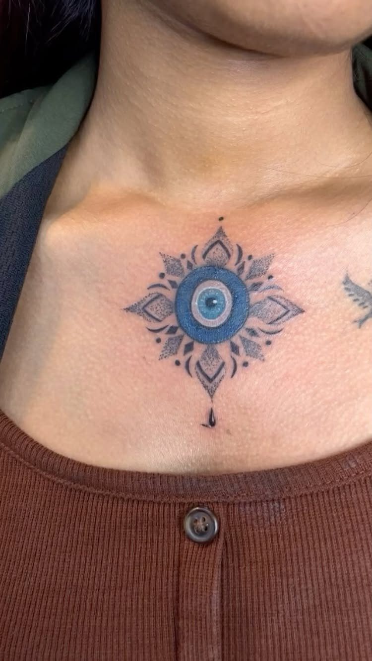 Evil eye tattoo with blue ink and mandala patterns on a person's chest.