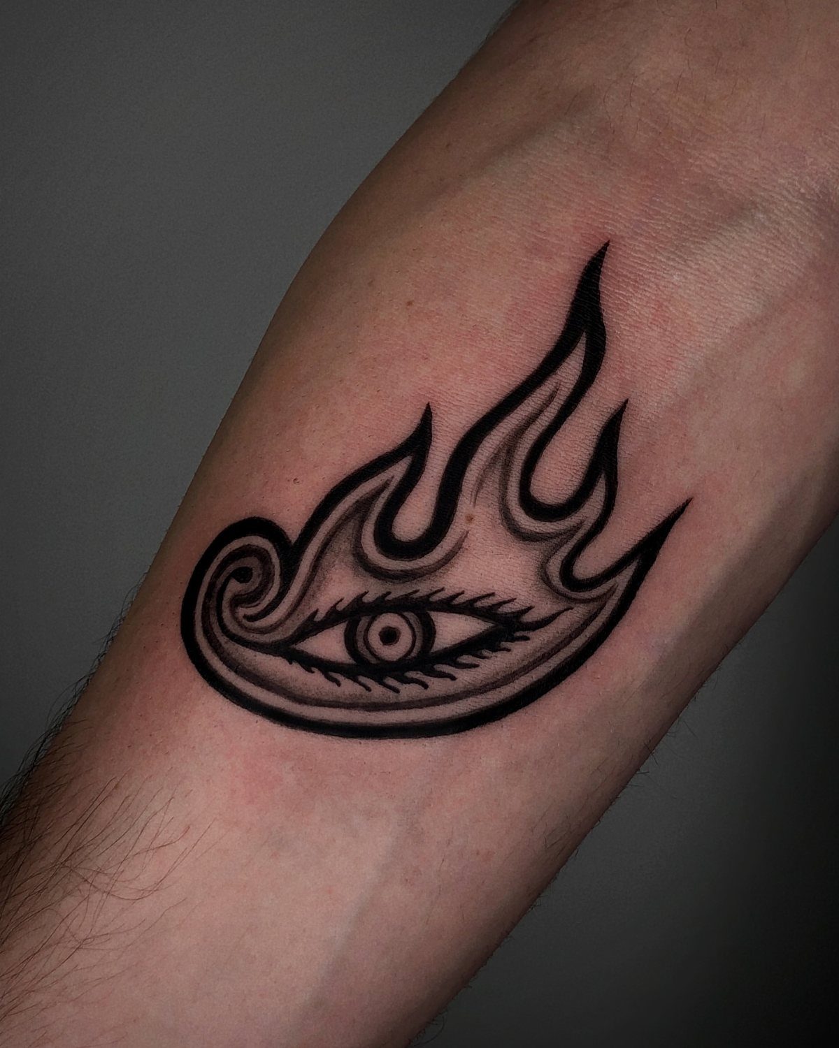Black ink tattoo of an eye inside a flame on a person's forearm.