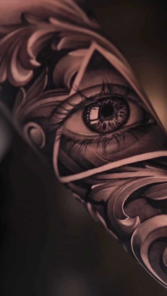 Realistic eye tattoo inside a triangle with ornate floral scrollwork on skin.