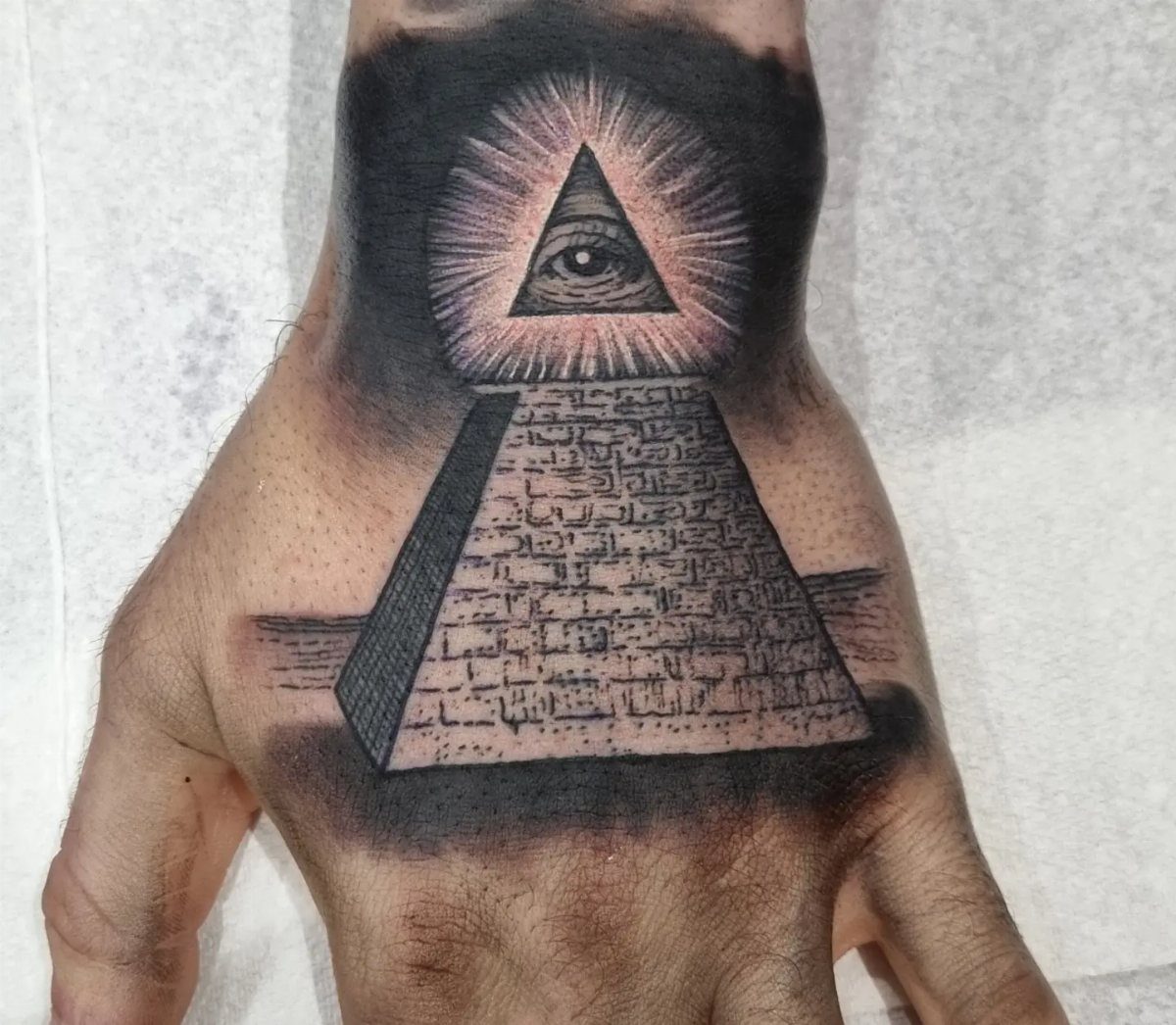 Pyramid tattoo on a hand with a third eye surrounded by radiating rays.