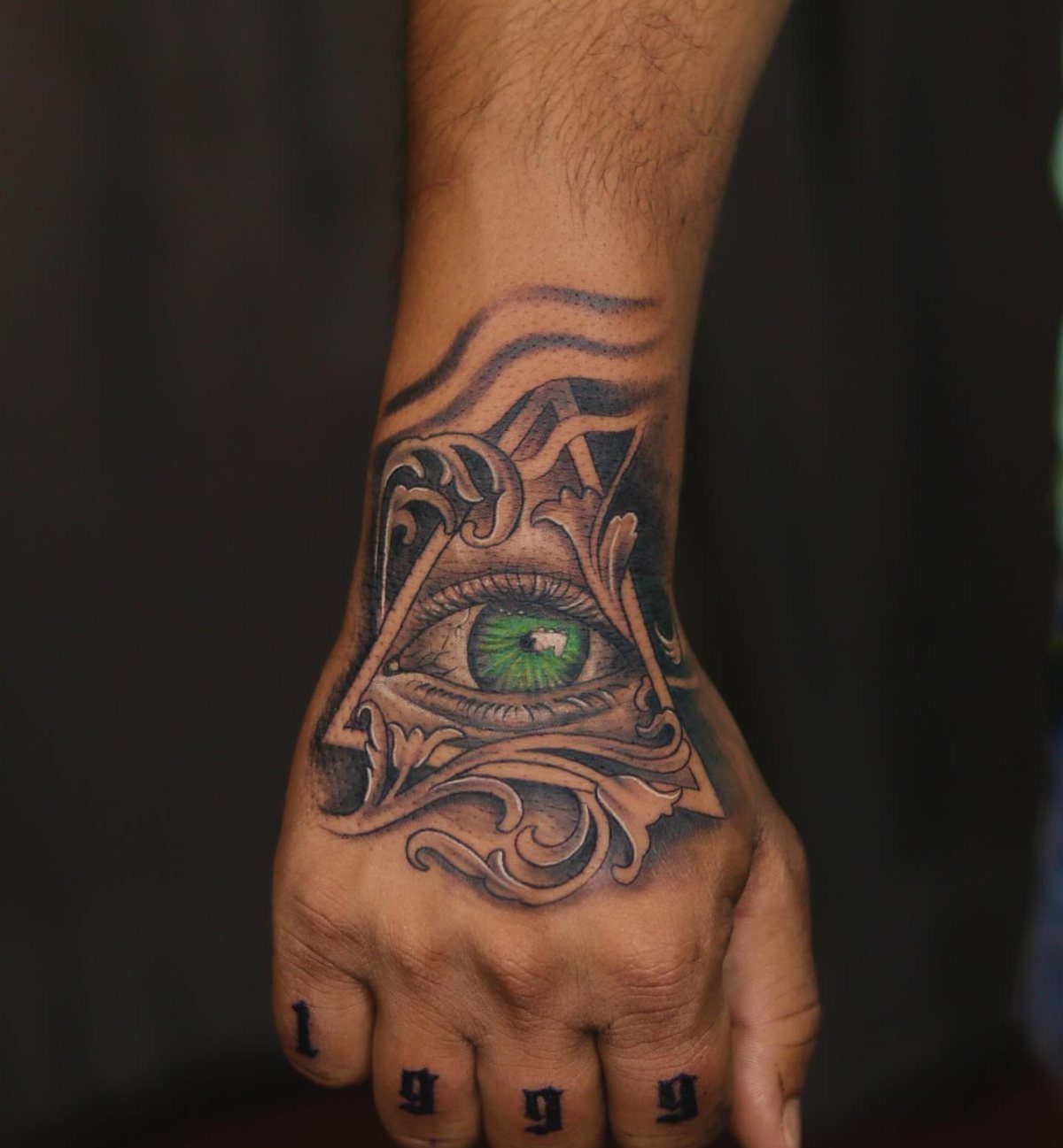 Realistic green eye tattoo inside a triangle with ornate grey filigree on a hand.