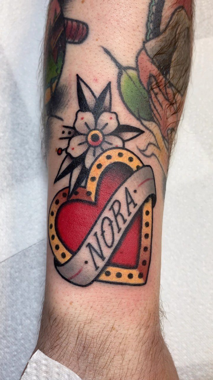 Traditional red heart tattoo with a banner reading NORA on a forearm.