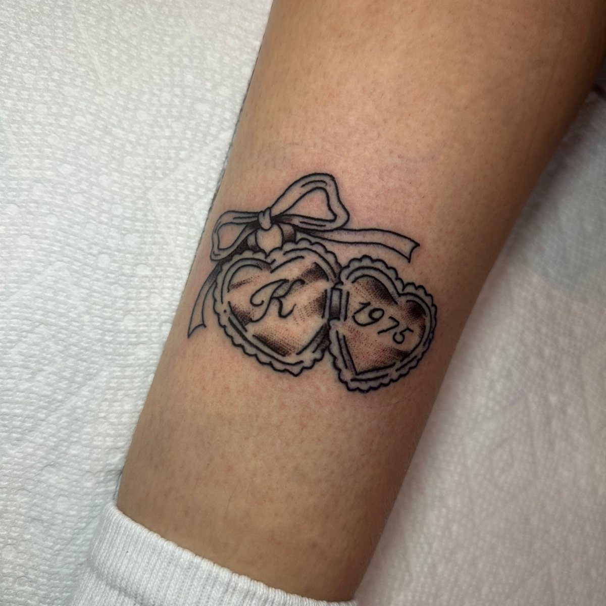Interlocking heart tattoos with a bow, the letter K, and the year 1975.