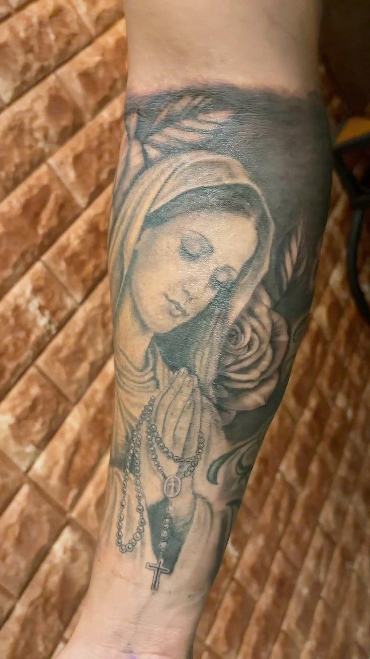 Black and grey tattoo of a praying woman and rose on a forearm.