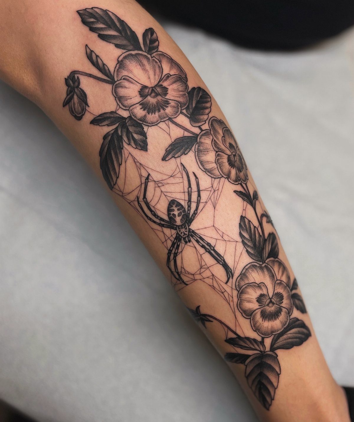 Spider web tattoo with pansy flowers on a person's forearm in black and grey