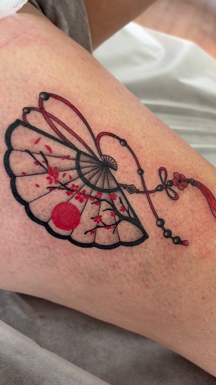 Tattoo of a Japanese folding fan with cherry blossoms in red and black ink.
