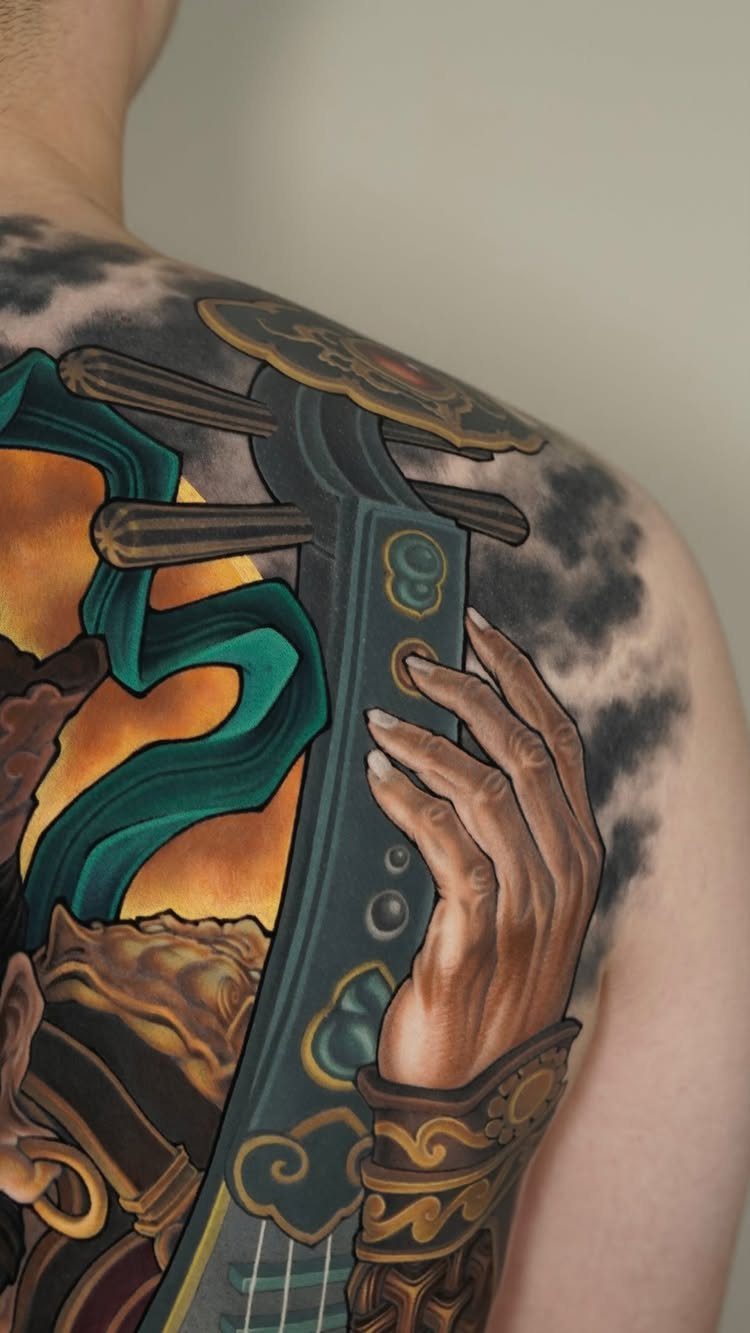 Neo-traditional tattoo on a shoulder depicting a hand holding a detailed musical instrument.