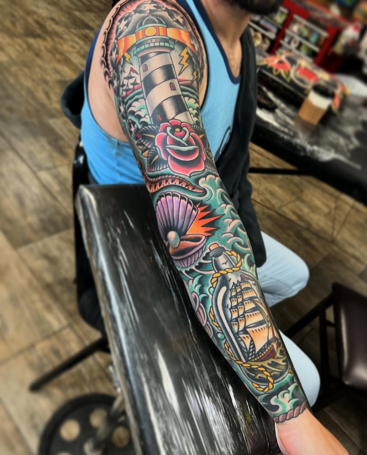 Traditional sleeve tattoo featuring a lighthouse, red rose, and ship in a bottle.