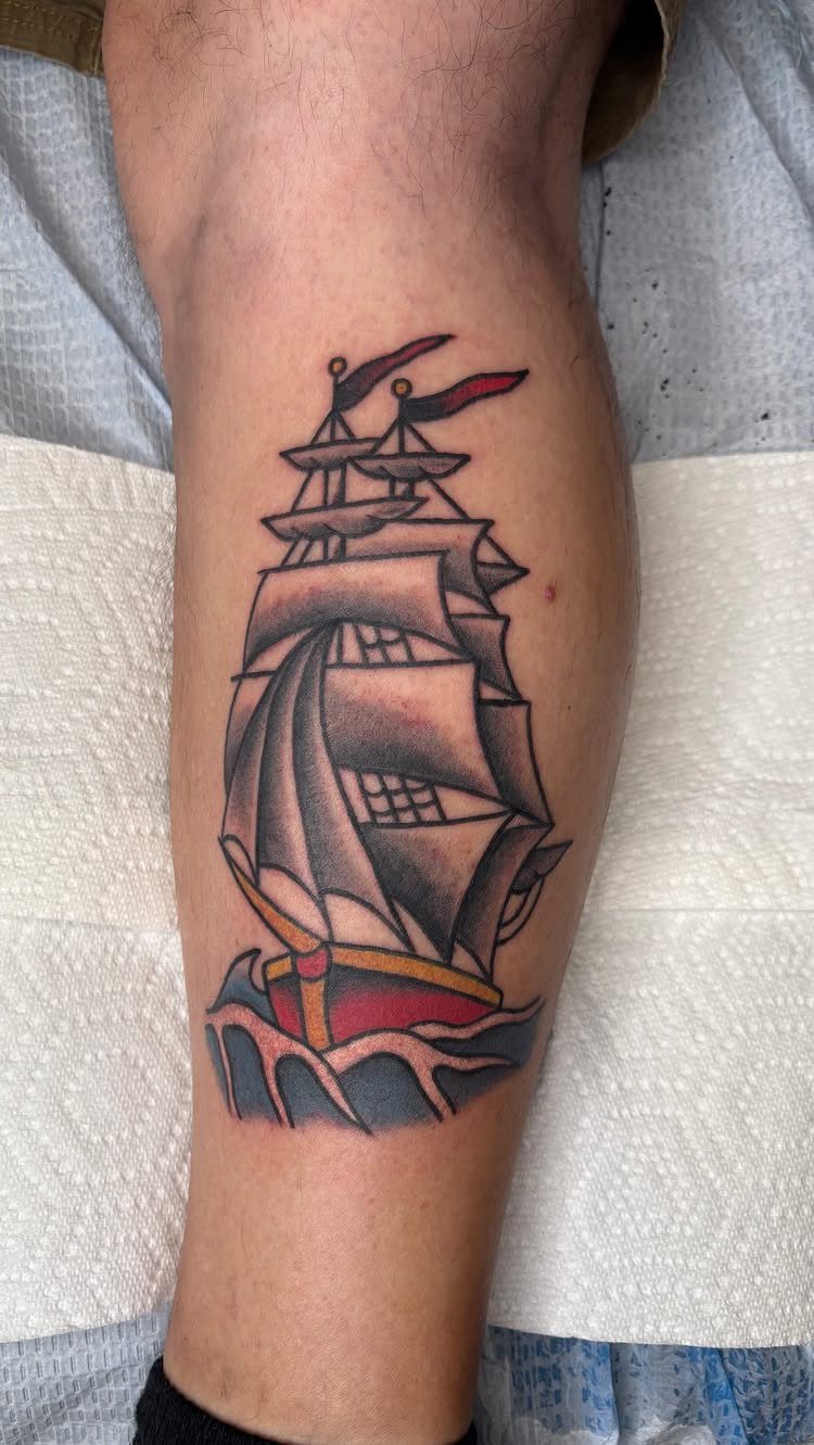Sailing ship tattoo with red hull and grey sails on a man's leg.