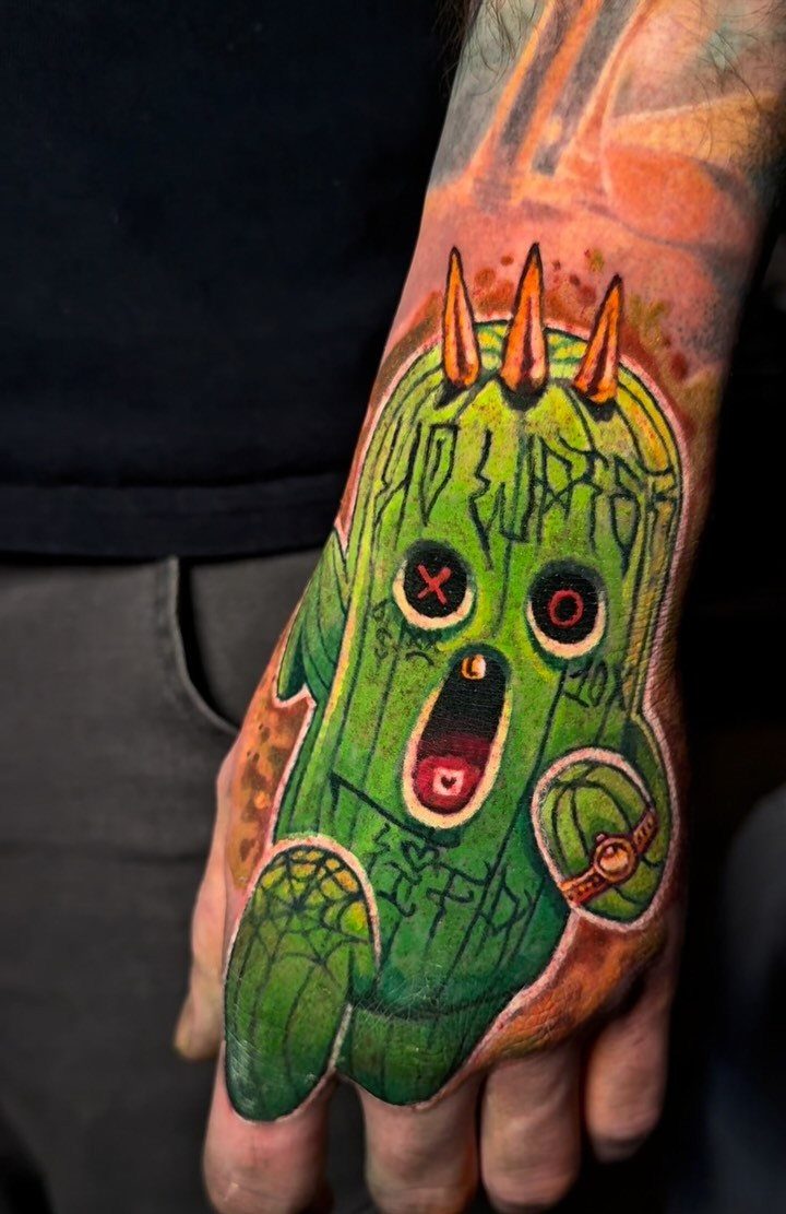 Green cactus character tattoo with yellow spikes on a person's hand.