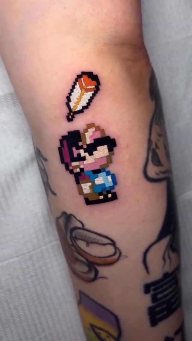 Pixel-art Mario tattoo on an arm with a floating orange feather above him.