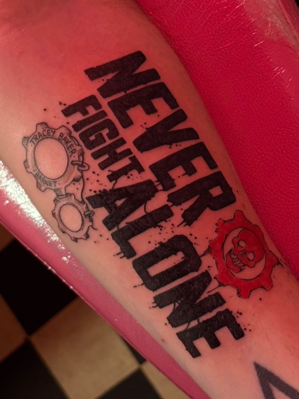 Tattoo on skin with the quote NEVER FIGHT ALONE and gear symbols.