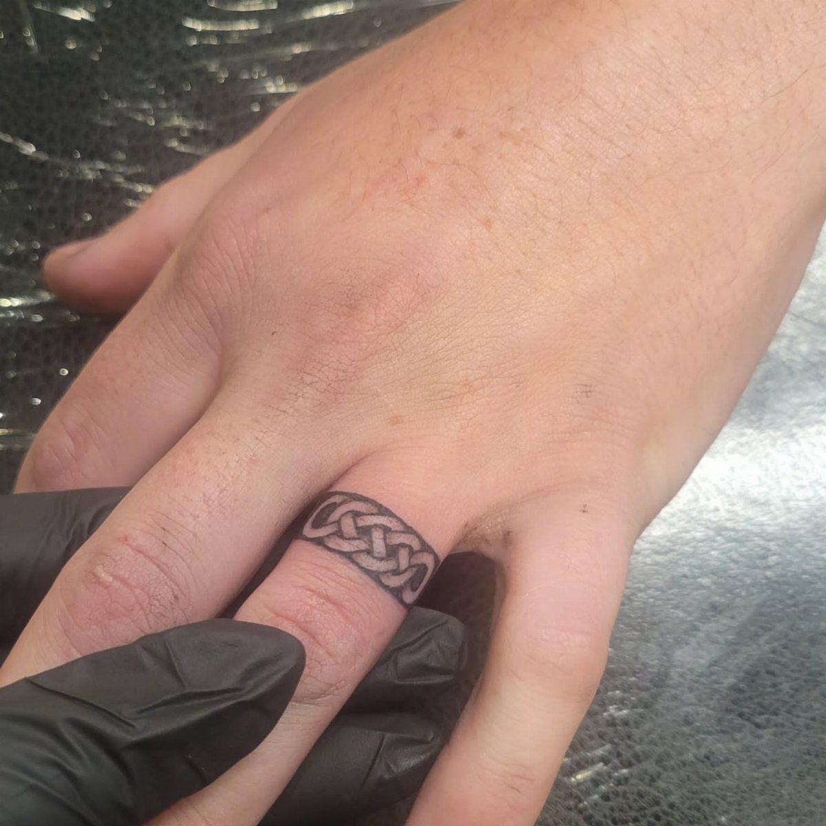 Celtic knot tattoo wrapping around a ring finger, held by a gloved hand.