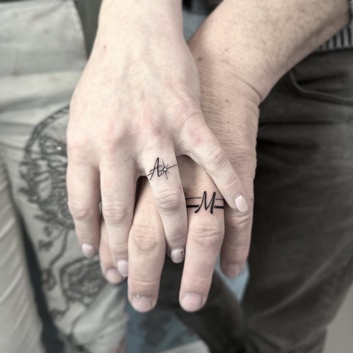 Two ring fingers with tattoos of the initials A and M in black ink.