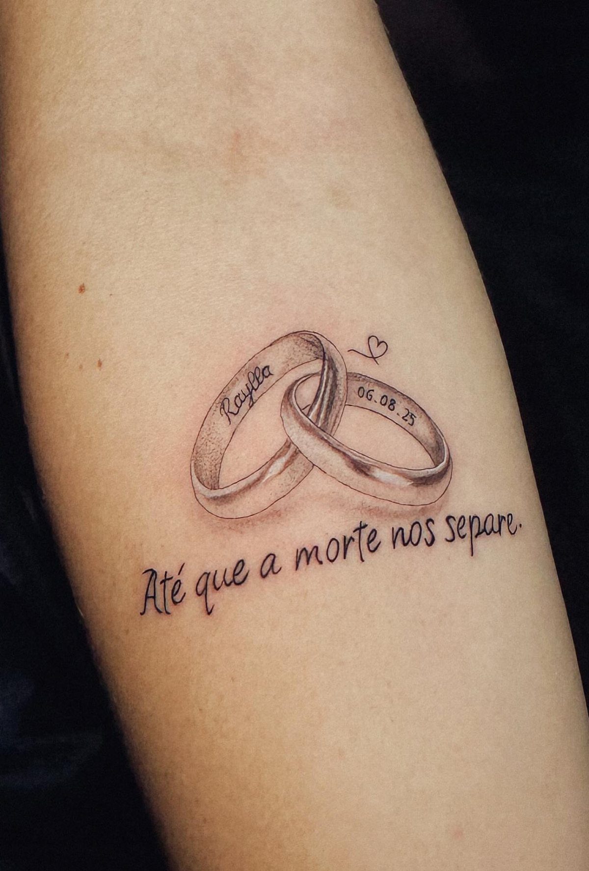 Interlocking wedding ring tattoos with a name, date, and Portuguese script on skin.