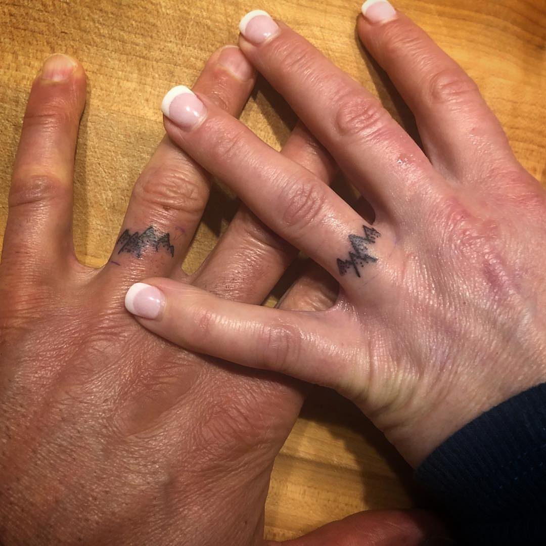 Two ring fingers with matching black mountain range tattoos on a wooden background.