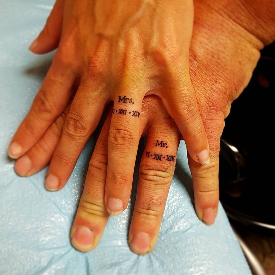 Two ring fingers with matching Mr. and Mrs. tattoos and Roman numerals