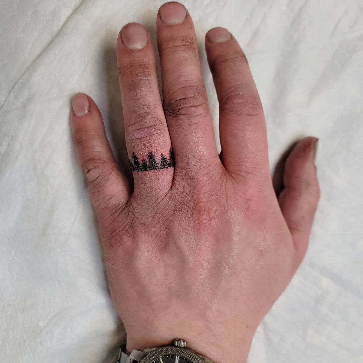 Hand with a black silhouette pine tree forest tattoo wrapping around the ring finger