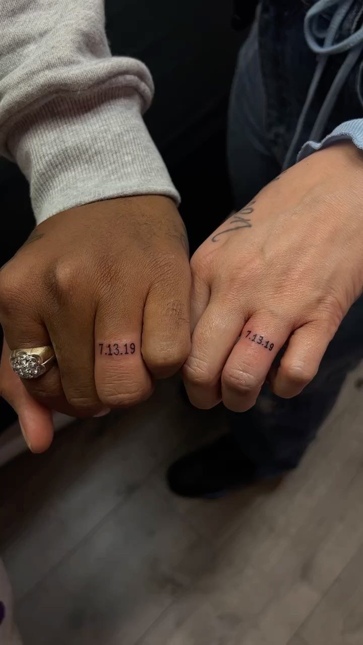 Two ring fingers with matching tattoos of the date 7.13.19 in black ink.