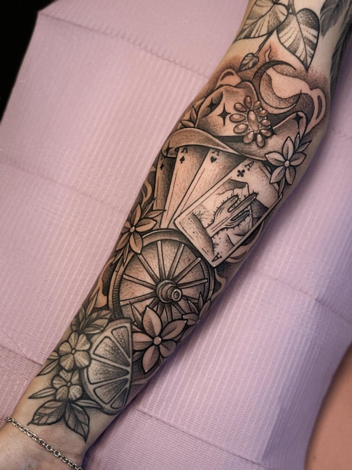 Black and grey tattoo of playing cards, cowboy hat, and wagon wheel on arm.
