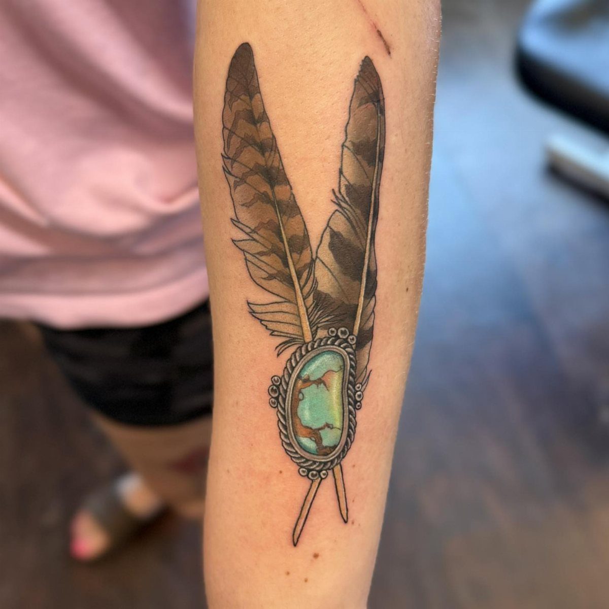 Tattoo of two brown feathers with a turquoise gemstone accent on an arm